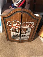 Closed wooden dartboard cabinet with Cheers logo and pub scene illustration on front closed doors.