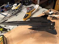 Die cast black model of SR-71A Blackbird on a wooden table with other model helicopters around. Shown from the side.