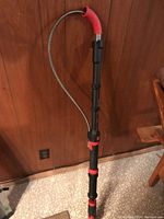 Full view of Milwaukee electric toilet snake standing upright against a wood-paneled wall, showing extended flexible metal cable and red handle grip.