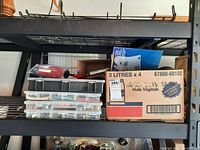 Black shelving with three transparent plastic bins stacked containing various electrical components. A cardboard box labeled '3 Litres x 4 Huile Vegetale' is also on the shelf, filled with other items.