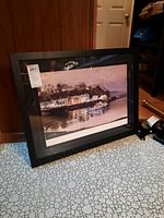 Framed print leaning against a wooden surface, showing the black frame and part of the floor.