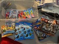 Photo showing Lego mini figure blind bags including Disney mini figure and Kinex Mario mini figures, along with their packaging inside a plastic bin.