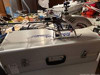 RC helicopter with visible rotors and servo components on metal hard case in background cluttered workspace.