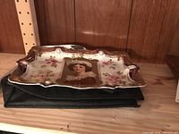 Front and side view of porcelain tray on black base showing overall shape, scalloped gilt border, ribbon/jewel motifs