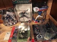 Five sports figurines from hockey, baseball, and football lined up on shelf showing full packaging and labels.