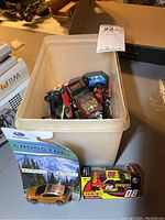 Plastic bin filled with various vintage toy cars showing wear, plus Subaru Crosstrek toy car and NASCAR Panini toy car box in front