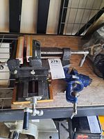 Two vises side by side on a wooden surface; one large bench vise with a wooden base and one smaller blue clamp on vise