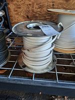 Metal spool holding white insulated electrical wire on a shelf with other similar spools. Wire end is frayed and spool is slightly rusty.