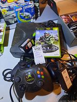 Original Xbox console with power adapter, three controllers, and Halo game placed on a table with price tag.