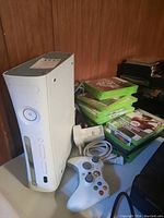 White Xbox 360 console next to a white wireless controller, power adapter, and a stack of Xbox 360 games in green cases.