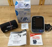 Photo showing the Omron automatic blood pressure monitor with cuff, original box, manuals, and power cord laid out on a wooden surface.