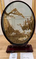 Front view of oval carved cork diorama framed in lacquered wood, depicting Asian landscape with cranes and pagoda buildings.