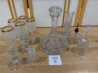 Full view of mixed lot on wooden surface including crystal decanter with stopper, 5 gold-rimmed wine glasses, two crystal dinner bells, two candle holders, and a small vase
