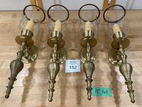 Four matching brass wall sconces with glass hurricane shades arranged side by side, showing overall set and size reference.
