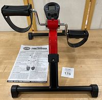 Complete view of the deluxe pedal exerciser with instruction manual placed behind it on a wooden table, showing red frame and black pedals with straps.
