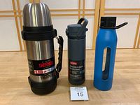Front view of three bottles: large Thermos with labels, smaller gray Thermos with label, blue Takeya bottle with rubber sleeve.