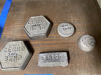 Photo showing six metal ingots on wooden surface including two octagonal Cerrobend ingots, two round ingots with CERO SAFE marking, small rectangular SAECO ingot, and a smaller round unmarked ingot.