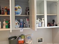 Photo of open cupboards showing various cleaning supplies including Tide laundry detergent container, Lysol spray, and smaller bottles of cleaning liquids.
