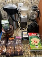 Overall lot lineup showing coffee pots, Braun coffee grinder, coffee bags, tea box, coffee filters, and accessories on a granite countertop.