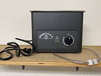 Side view of Grobet USA ultrasonic cleaner showing power cord and transparent drain tube with clamp, metal body with stainless steel rim.