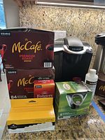 Black Keurig coffee maker with metallic silver lid, alongside boxed coffee pods McCafe Premium Roast Medium, Folgers Black Silk, Twinings green tea pods, Keurig cleaning solution bottle and water filter cartridge box on countertop.