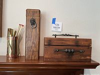 Photo showing the three items on a wooden surface against a wall: brass match holder with long matches, tall wooden box with lion head ornament, wooden box with drawer and metal handles