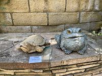 Photo showing both turtle and frog statues on stone patio against a brick wall. Turtle is tan with detailed shell, frog is gray with textured skin.