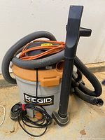 Front view of Ridgid ShopVac showing attached hose, extension cord wrapped around top, vacuum attachments leaning against unit, and orange bottom drain valve.