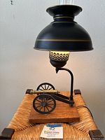 Full view of the vintage lamp showing black metal shade, lit bulb, and miniature cannon on wooden base.