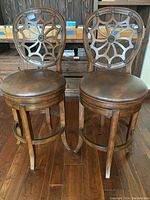Front view of the two dark wood and dark brown leather swivel bar stools side by side on wooden floor showing seats and intricately carved backrests.