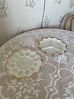 Two white milk glass plates on lace tablecloth. One round egg plate with gold trim, one 3-section divided plate with gold trim.
