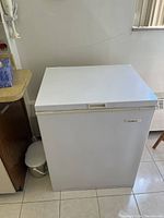 White Beau Mark deep freeze chest freezer, closed lid, placing on tiled floor near radiator and wooden cabinet.
