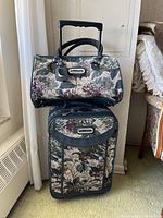 Vintage floral print suitcase with extendable handle and matching duffel bag stacked on top. Both items made of tapestry-style fabric with leather trim and Oscar de la Renta logo plaques visible.