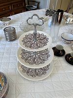Full assembled 3-tier ceramic serving tray with black and white intricate pattern, displayed on a white quilted surface with various other tableware items around.