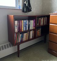Front view of vintage mid-century modern wooden bookcase filled with books, resting on two front legs and leaning on radiator at back