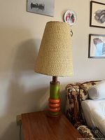 Full view of the Mid-Century Modern lamp placed on a wooden side table, showing green ceramic body with orange rings and tall woven beige lampshade in conical form.
