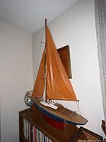 Front-side angled view of the model sailboat showing orange sails, blue and red painted hull, and wooden stand.