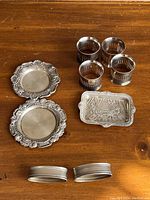 Overview of all six silver napkin rings, two ornate miniature round silver plates, and one rectangular miniature silver plate with detailed scene to show the complete lot.