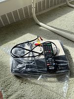 Sony Blu-ray DVD player with remote control and AV cables resting on carpet, inside a plastic bag.