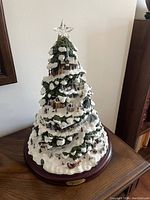 Full view of Thomas Kinkade Village Christmas illuminated tree figurine, 15 inches tall, showing snow-covered pine branches and village elements underneath.