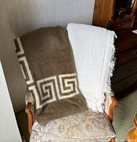 Two blankets draped over an upholstered patterned chair, one brown with geometric design and one white with fringed edges