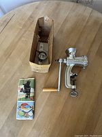Full view of metal meat grinder with original box and instruction pamphlet on wooden table.