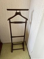 Wooden suit stand placed against a wall with visible hanger and bars for clothes.