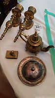 Photo of all lot items: two brass candlestick holders, brass teapot, silver-plated saucer with lid, and brass Chinese dog matchbook holder.