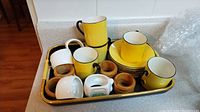 Full set includes yellow demitasse cups with black handles, yellow saucers, wooden napkin rings, white ceramic napkin holders, and a ceramic container on black and gold tray.