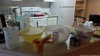 Kitchen counter displays all lot items: Fire King bowl, Tupperware bowl with lid, plastic measuring cups, silicone measuring cups, Corningware sauce maker and nested cookie cutters.