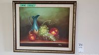 Full view of the framed oil painting showing a blue jug, grapes in a basket, and apples with a dark background.