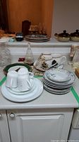 Photo shows assorted dinnerware including cups, plates, bowls, and a glass Christmas tree decoration arranged on kitchen counter.