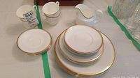 Overview of tea/coffee cups, creamer with plate, and stacked plates showing gold trim and white color.