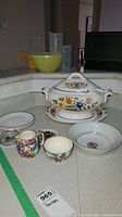 Full lot including soup tureen, Royal Winton cream and sugar set, plates, bowl, and additional yellow mixing bowl with measuring cups in background.